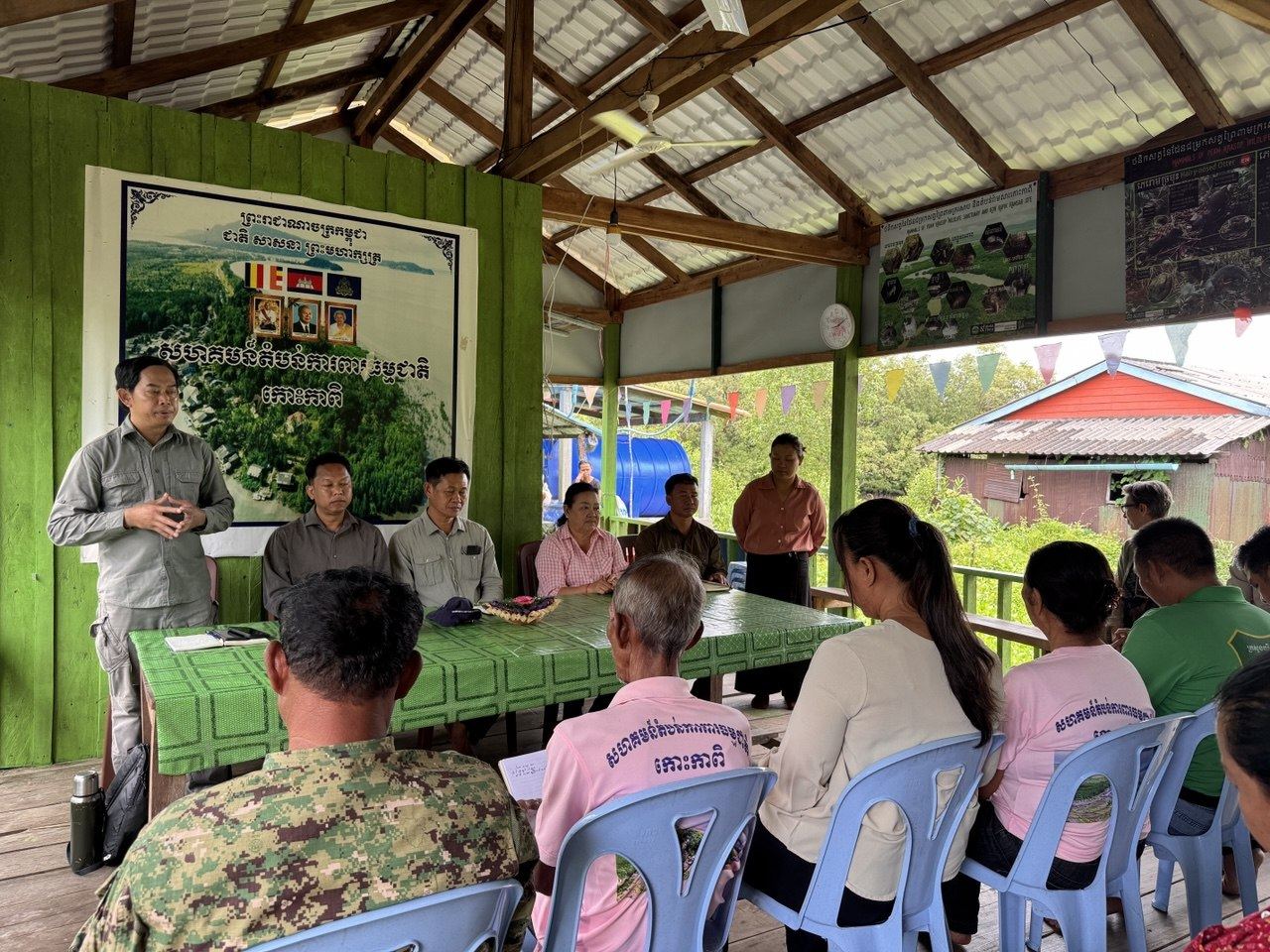 Community members seated in an open wooden pavilion listening to a facilitator