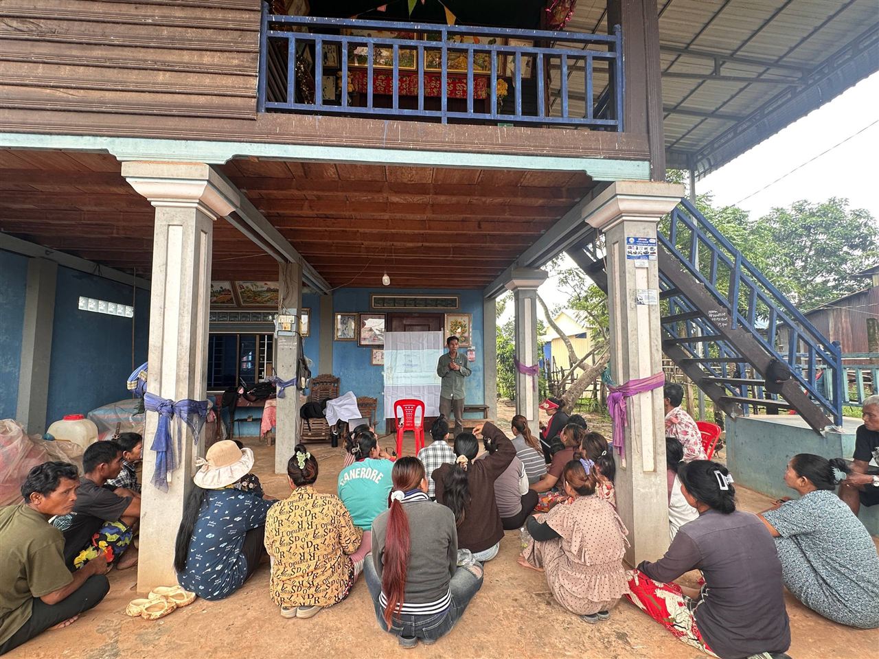 A facilitator presents to waste-picker household members seated on the ground beneath a raised wooden house