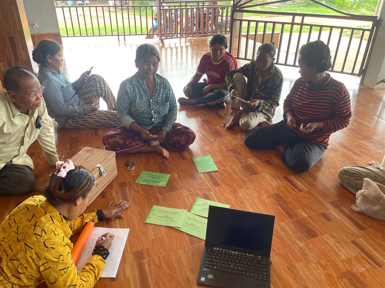 A group member records contributions in a ledger beside a savings box and green passbooks