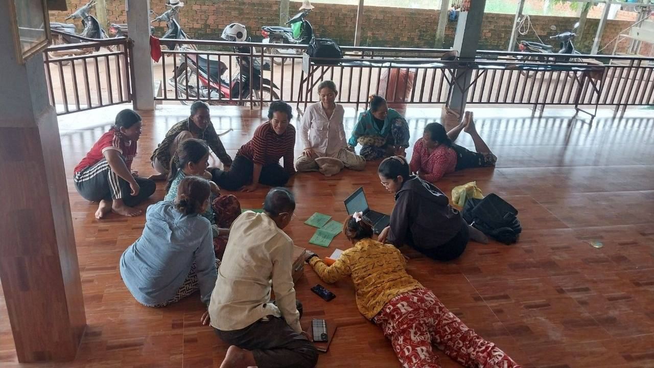 Women seated in a circle on a wooden floor with savings books and a laptop during a group meeting
